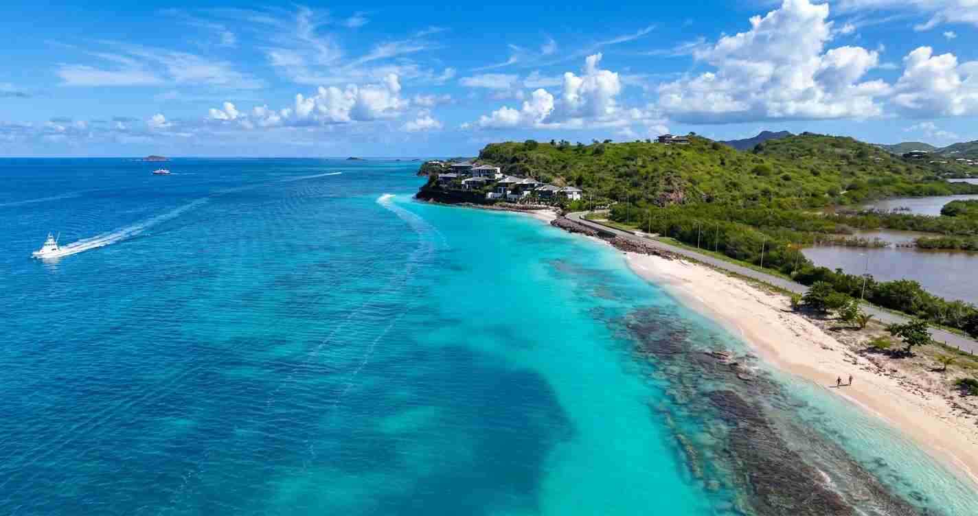 Aerial view of a tropical coastline: turquoise water, white-sand beach, green hills, and boats offshore.