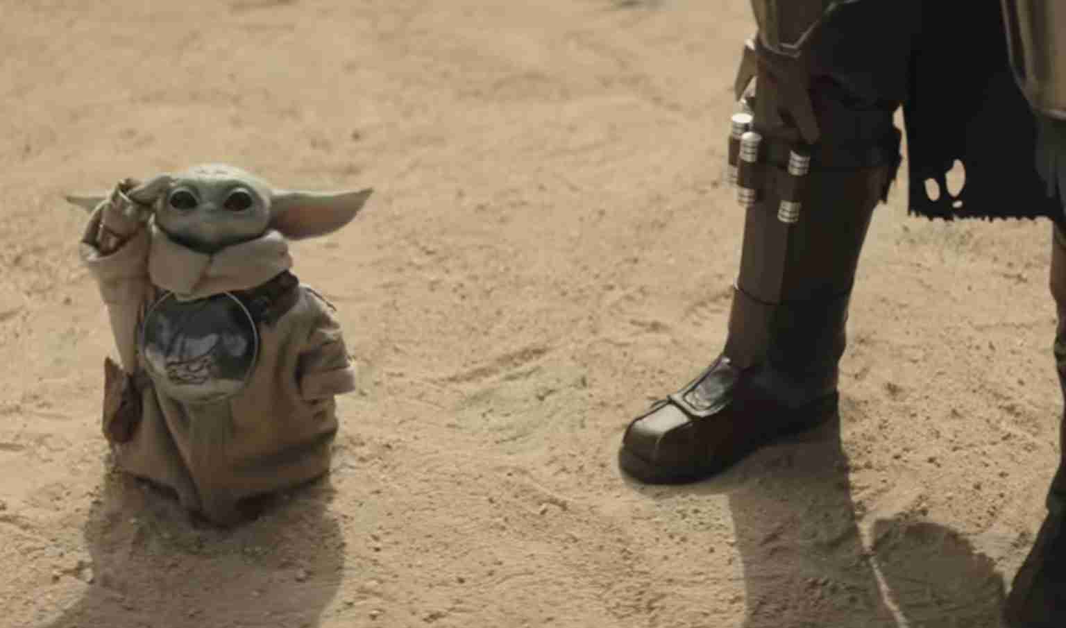 Grogu (Baby Yoda) in a brown robe stands in a sandy desert beside a armored figure's booted leg.