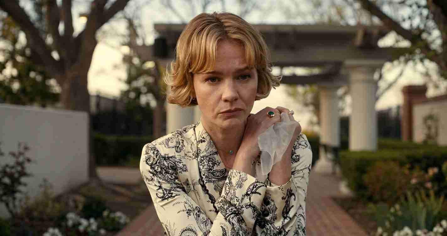 Woman with short blonde hair in a patterned blouse stands outdoors, holding a tissue and looking solemn.