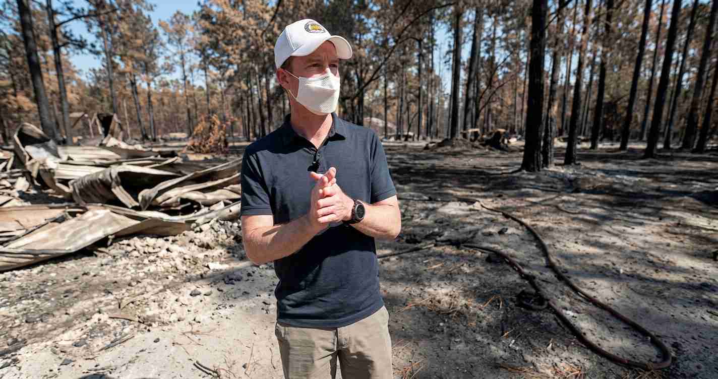 Man wearing a face mask and white cap stands in a burned forest with charred ground and debris in the background.