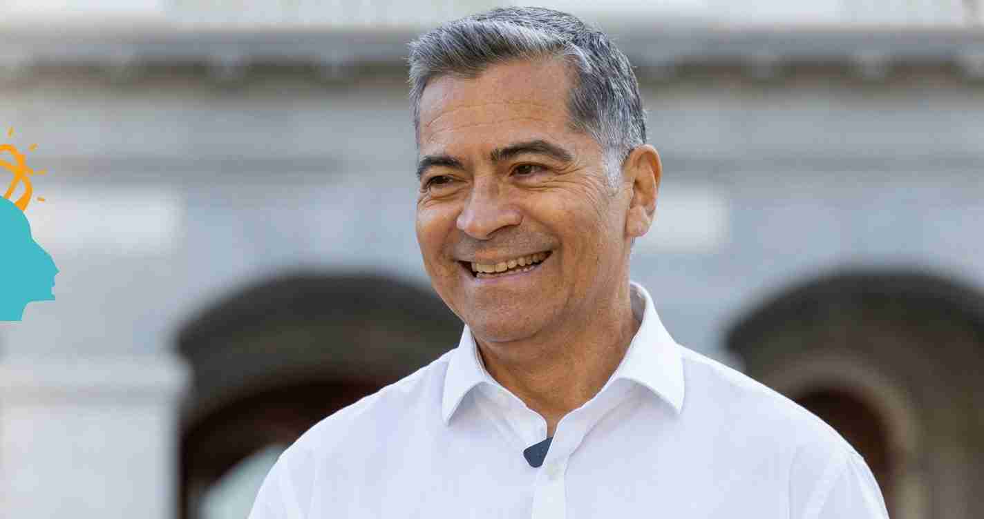 Smiling middle-aged man with gray hair in a white button-down shirt, outdoors with a soft background.