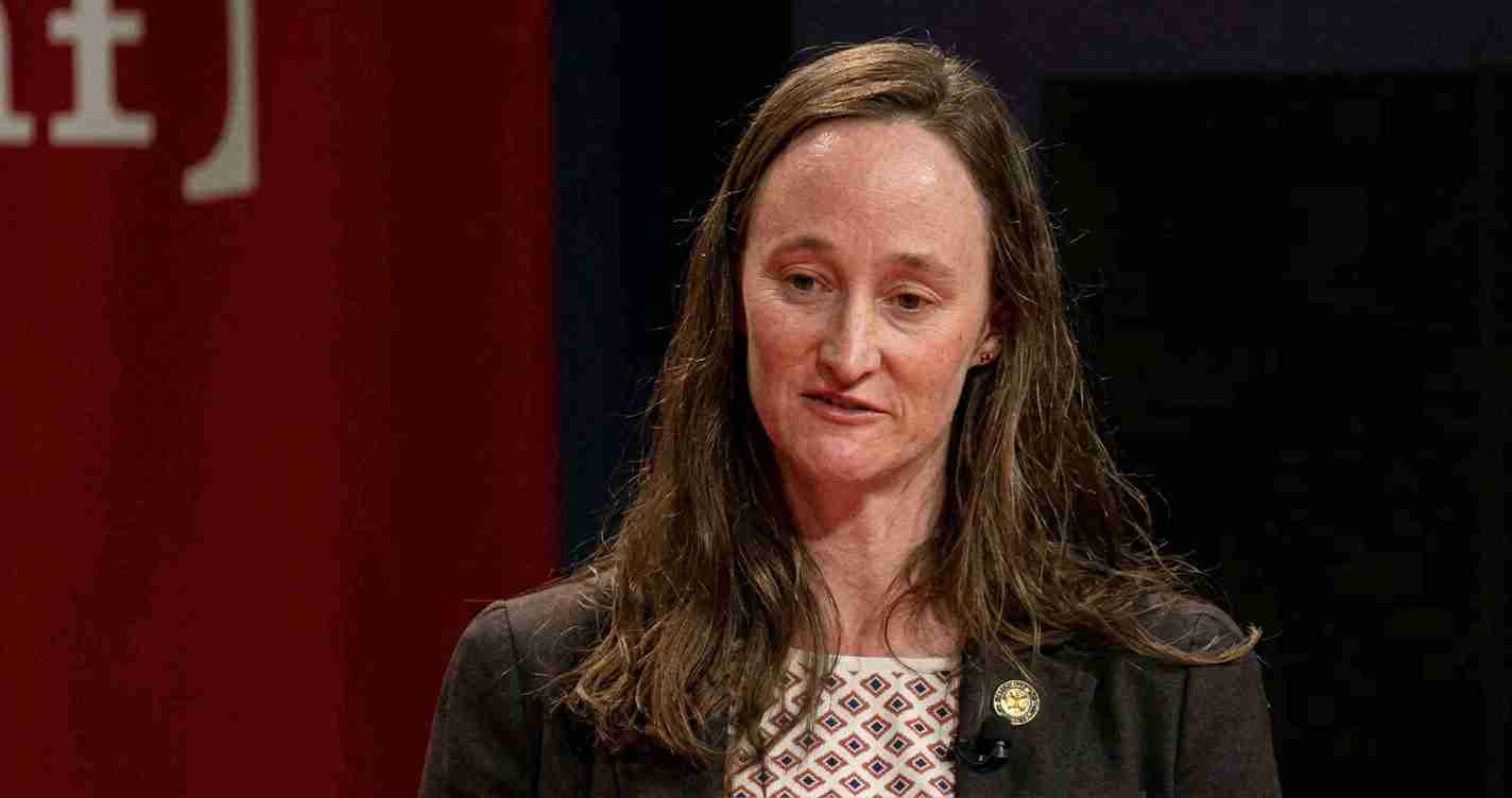 Woman with long brown hair in a dark blazer and patterned blouse speaking on stage, wearing a round lapel pin