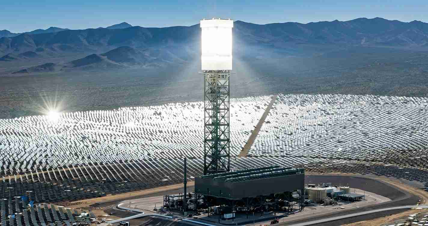 Solar thermal power plant with a tall central tower surrounded by rows of mirrors in a desert valley, mountains in the distance.