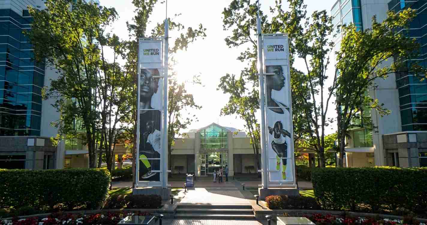 Entrance to a campus building framed by two tall banners that read 'UNITED WE RUN,' with trees and a sunny walkway and people near the doors.