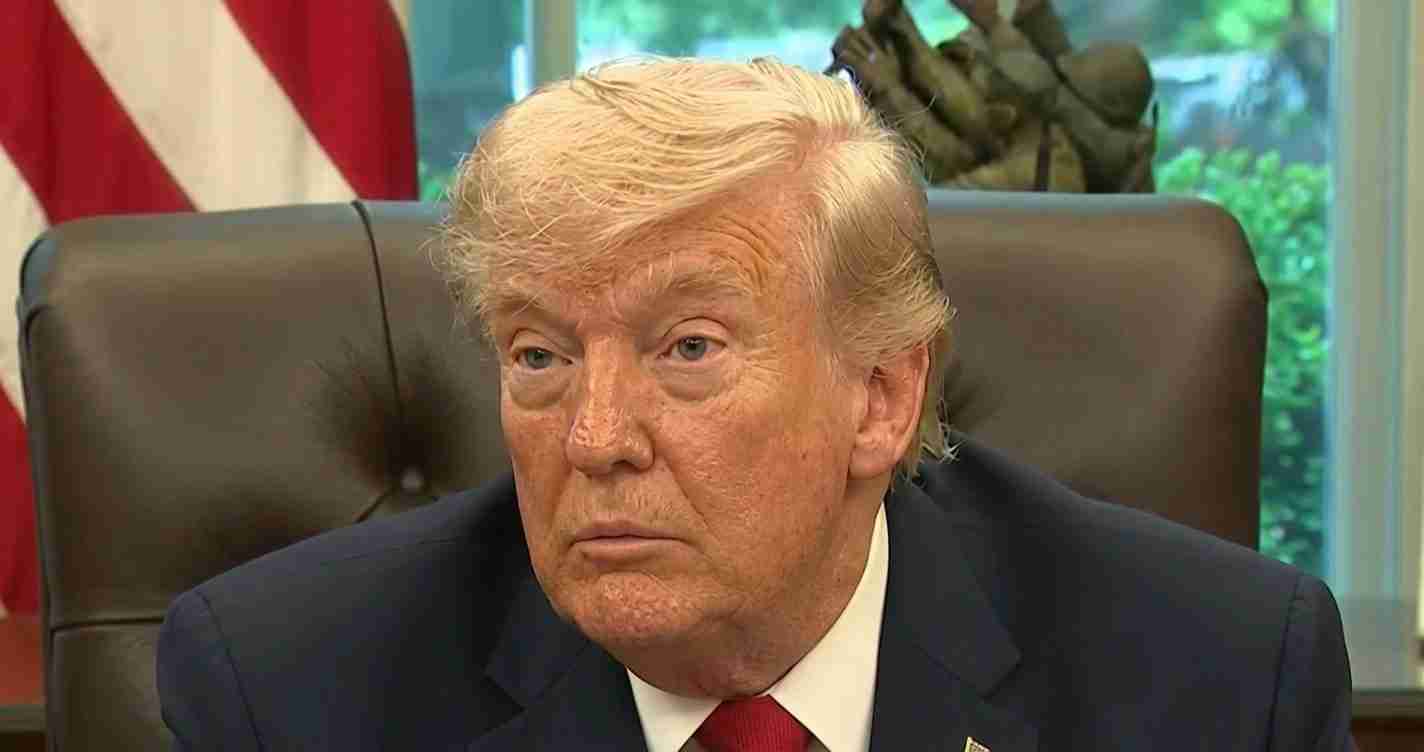 Close-up of a man with light blond hair in a dark suit and red tie, seated at a desk with an American flag in the background.