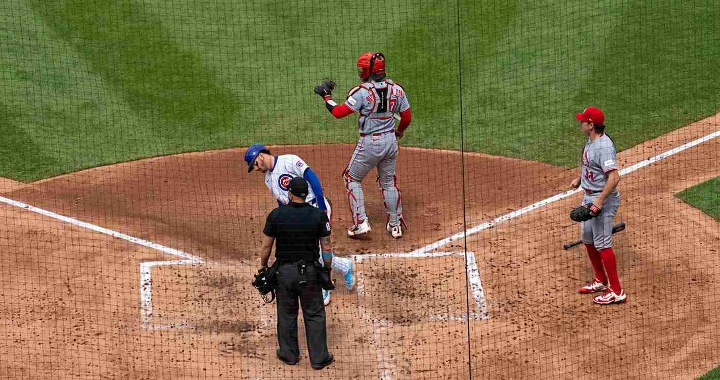Baseball scene at home plate: an umpire stands behind the batter as a White-Blue team player in a blue helmet prepares, with a gray-uniform catcher nearby and another gray-uniform fielder to the right.