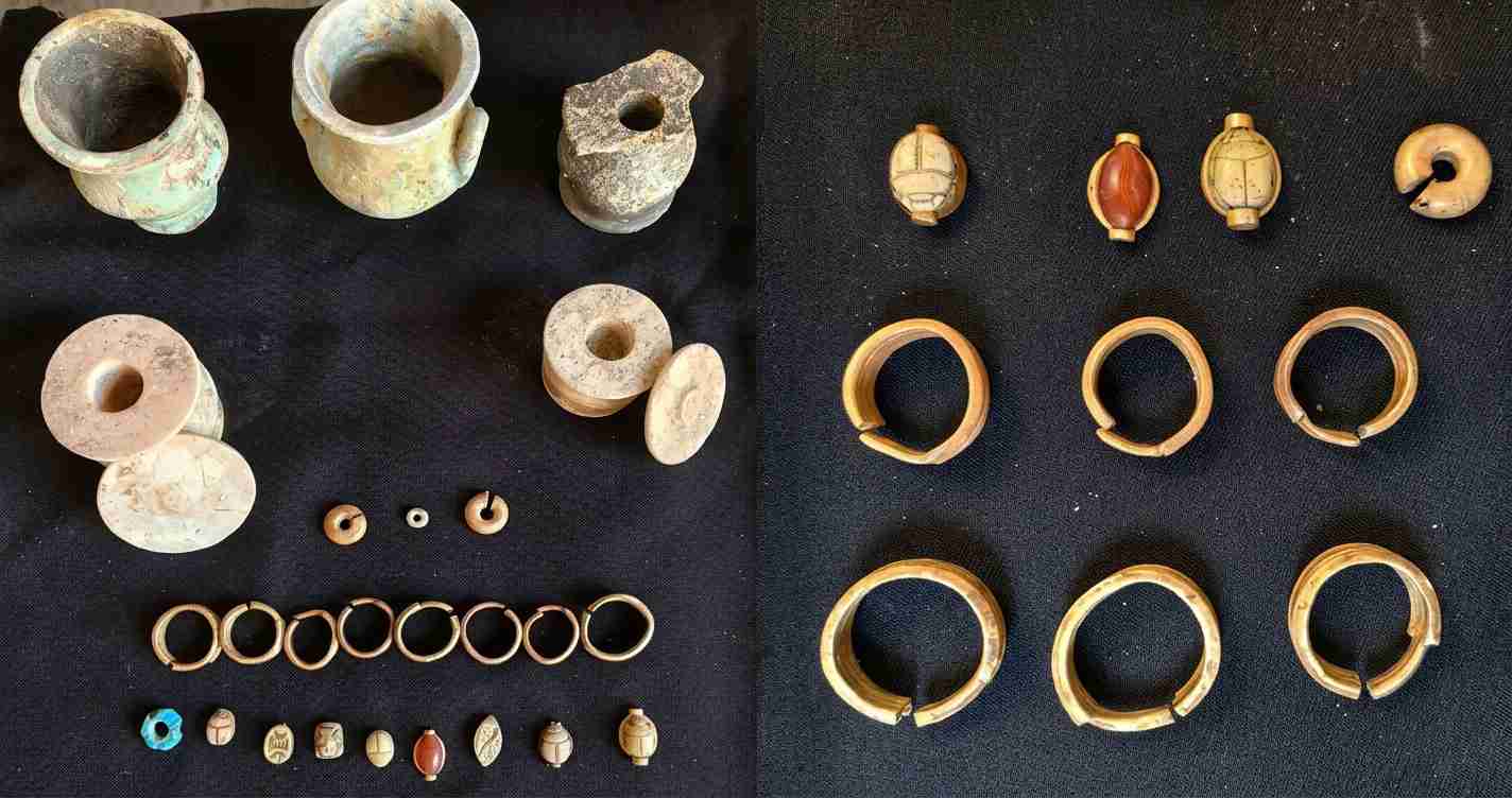 Display of ancient jewelry and pottery: clay cups, spindle whorls, and a row of small carved beads beside circular bangles on black fabric.