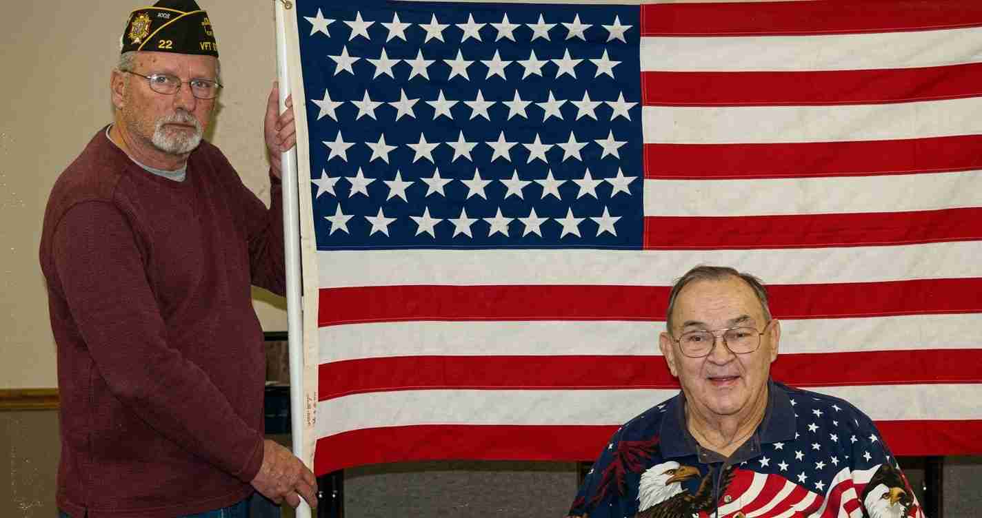 Two older men pose with a large American flag; one wears a veterans cap and maroon sweater, the other sits in a star-spangled shirt and smiles.