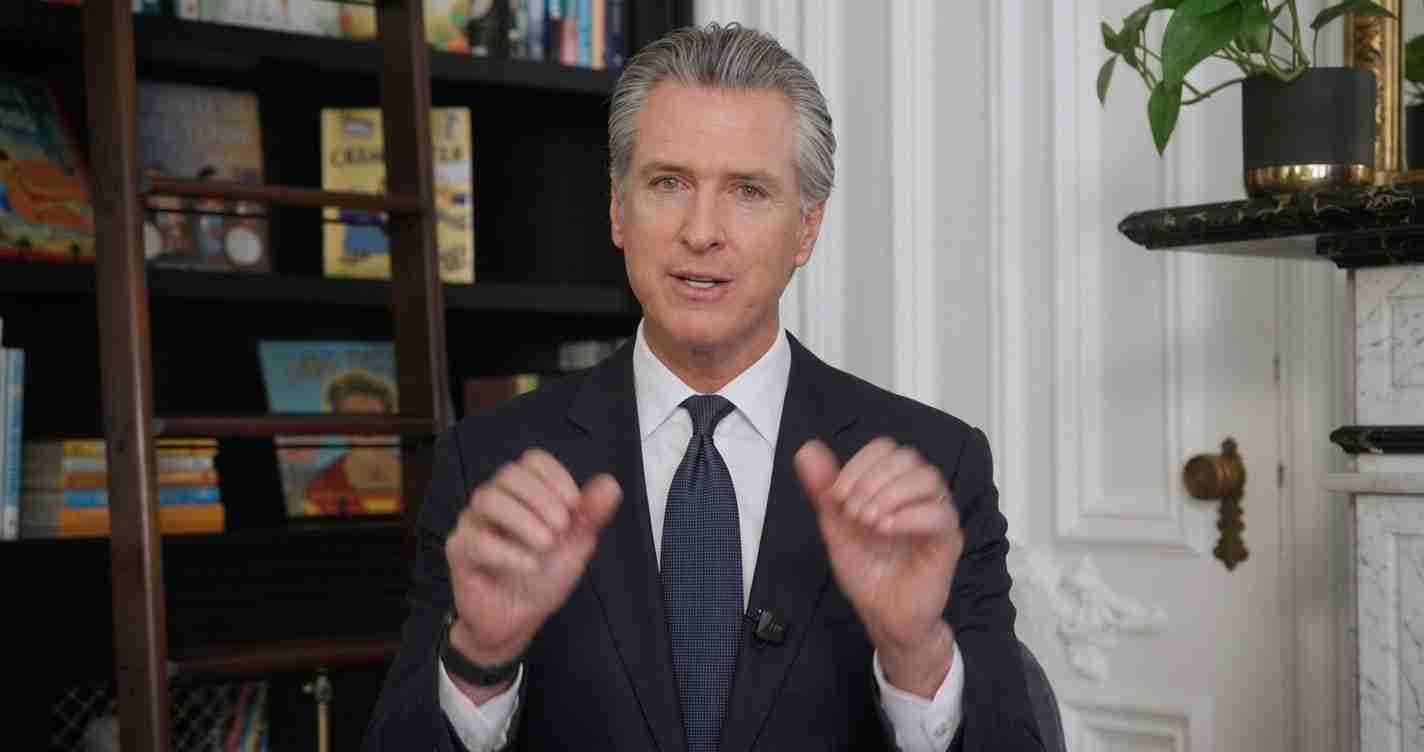 Man in a dark suit and tie sits in a study, gesturing with his hands while speaking.