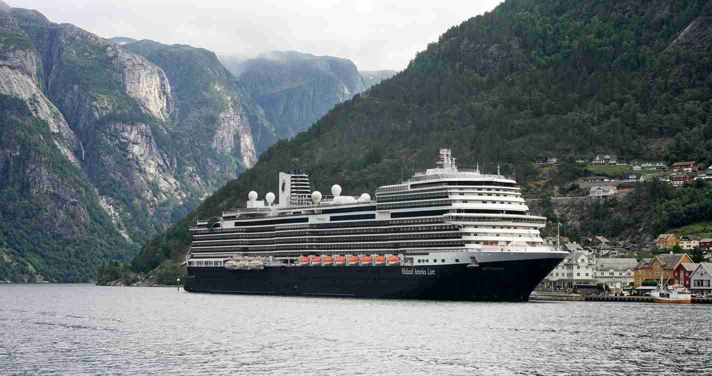 Large cruise ship anchored in a fjord with steep green mountains and a small coastal town nearby.