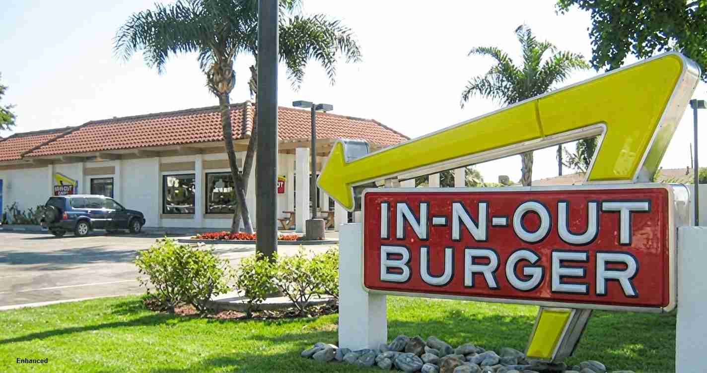 Bright In‑N‑Out Burger sign with a large yellow arrow, storefront and palm trees in the background.