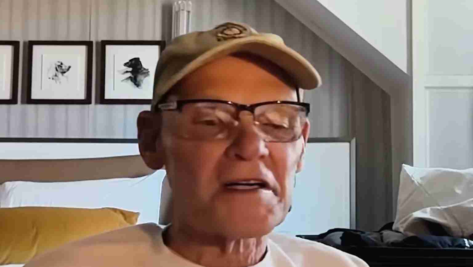 Older man wearing a beige cap and glasses speaks, seated in a bedroom with framed dog pictures on the wall behind him.
