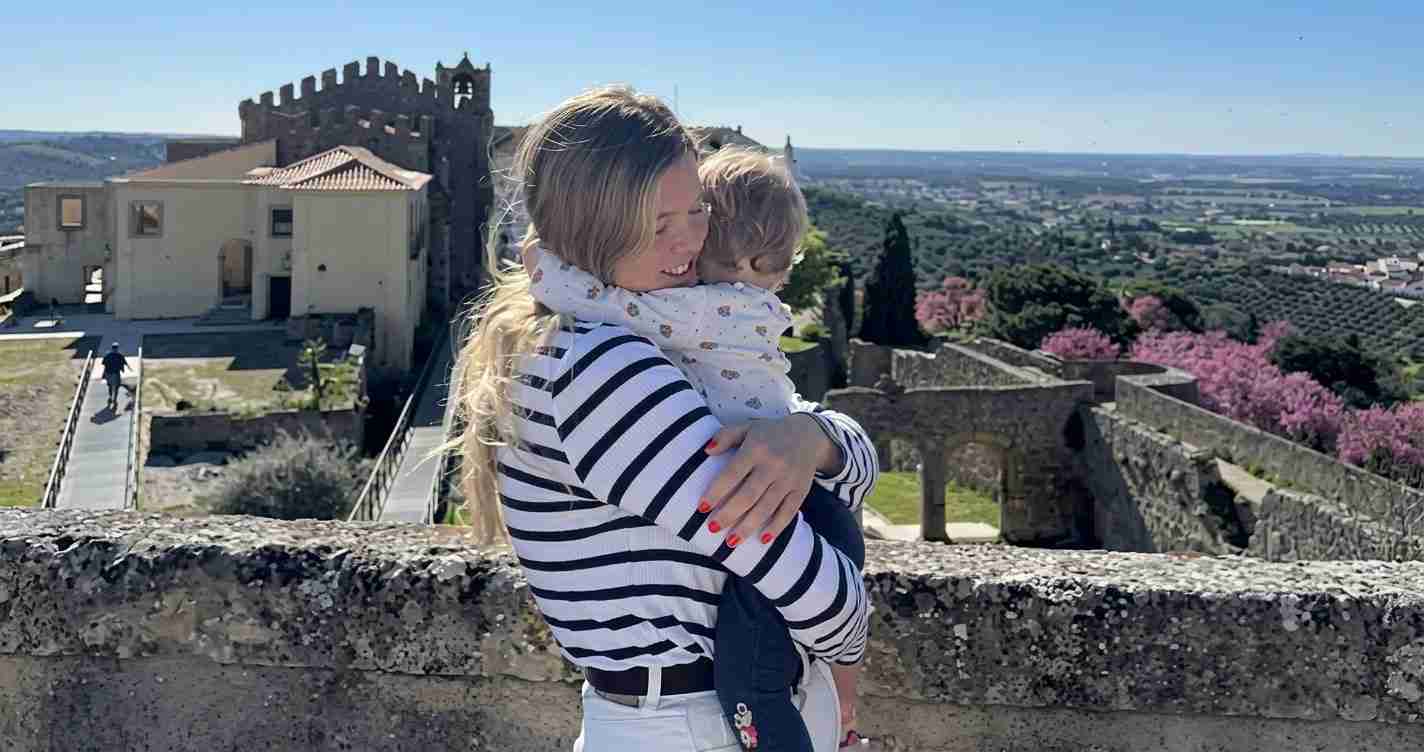 Woman in a striped shirt hugs a child against a stone wall with a castle and valley in the background.