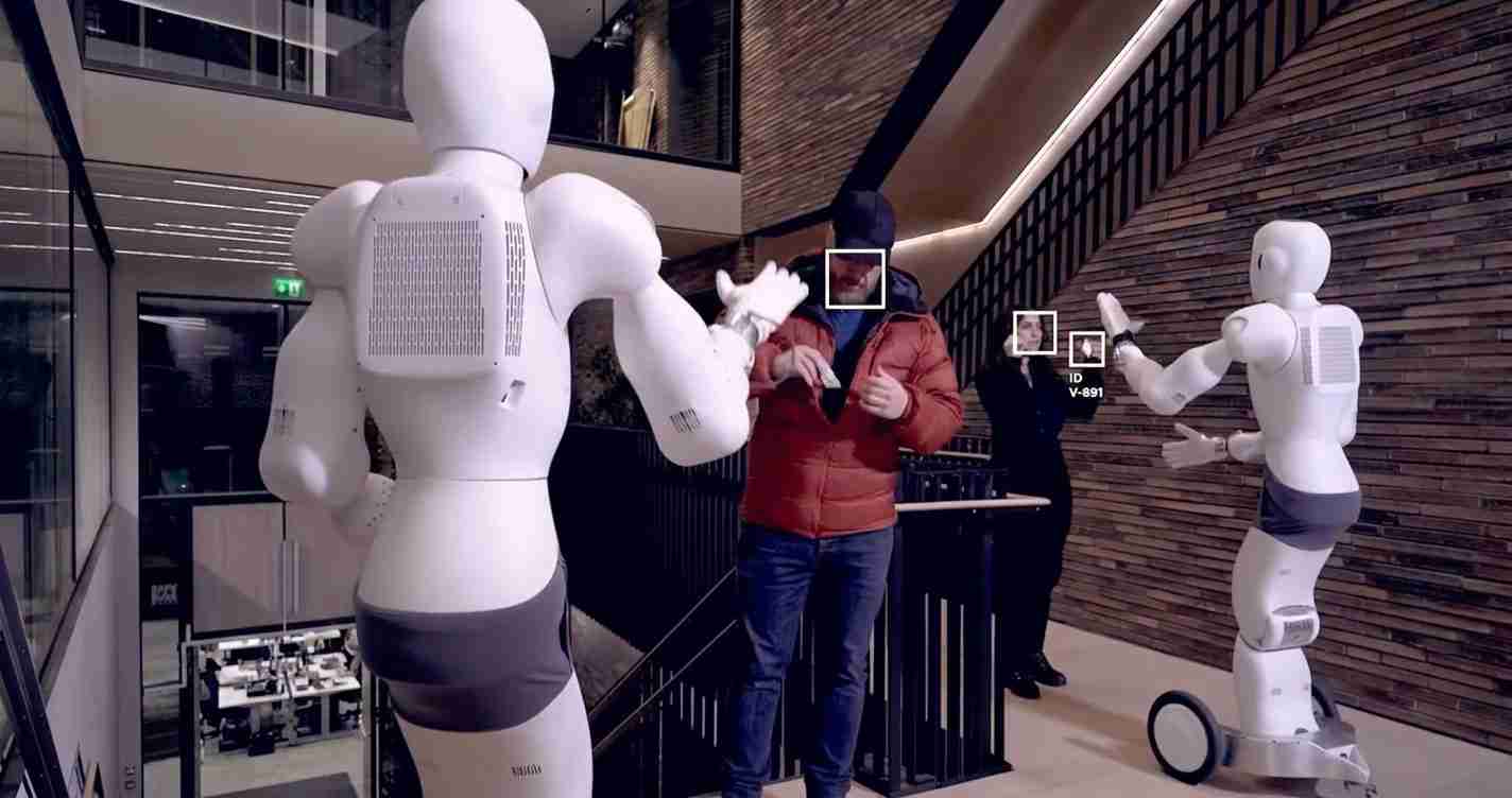 Two white humanoid robots stand on either side of a man in a red jacket, as they gesture in a modern indoor space. concise display of a robotics demo in a sleek building.