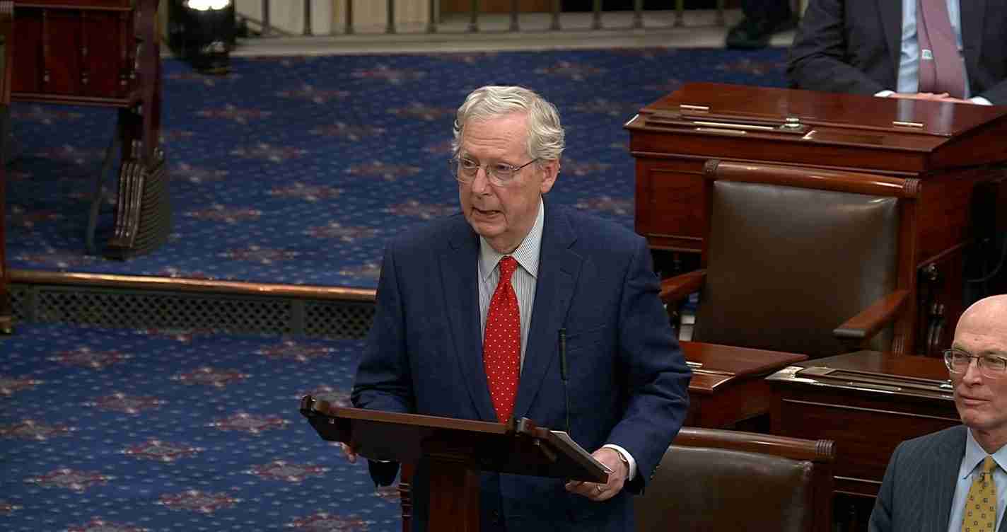 Man in a navy suit and red tie speaks from a wooden lectern on a blue patterned Senate floor, with another suited person seated nearby.