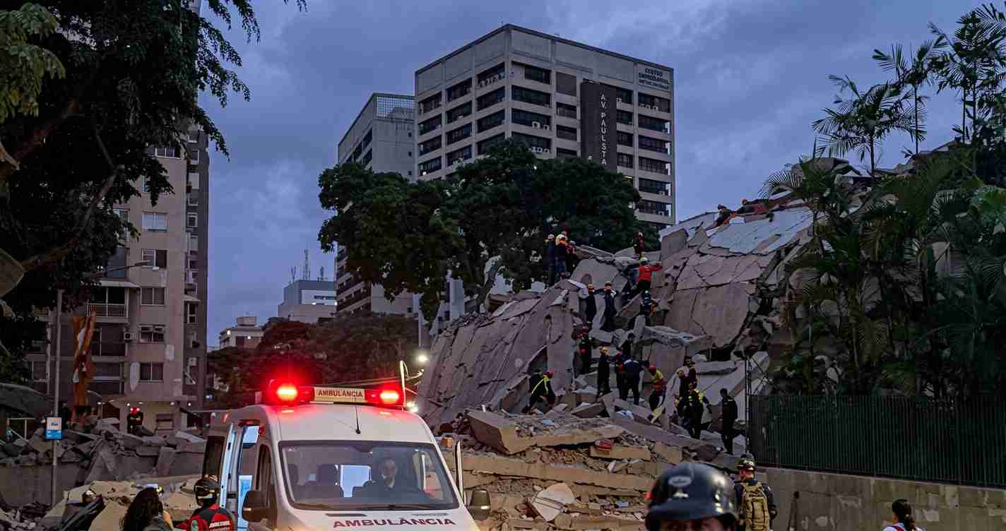 Rescue workers search a collapsed concrete building with an ambulance in the foreground at dusk.