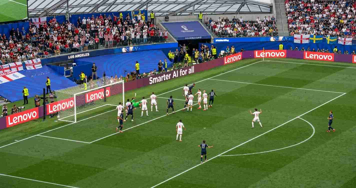 Soccer match near the goal with players in white and dark kits clustered by the net as a ball approaches, Stadium crowd visible in stands and advertising boards along the sidelines.