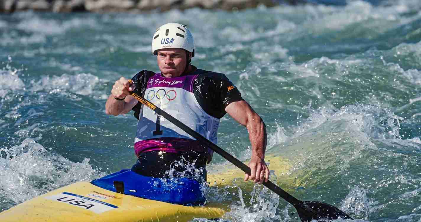 Male kayaker wearing a white helmet and USA jersey paddles a yellow kayak through rough whitewater.