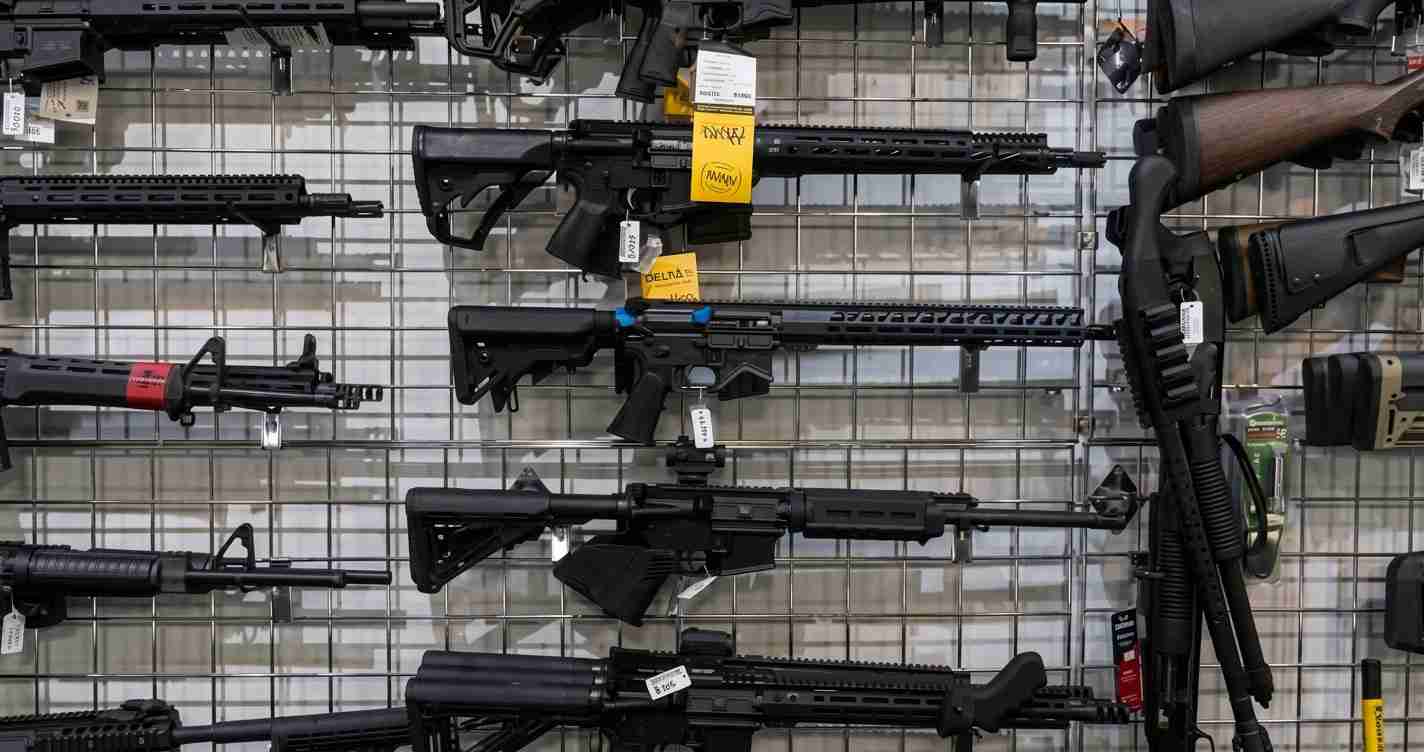 Row of black semi-automatic rifles mounted on a grid wall in a gun shop, each with price tags and accessories visible in the display.