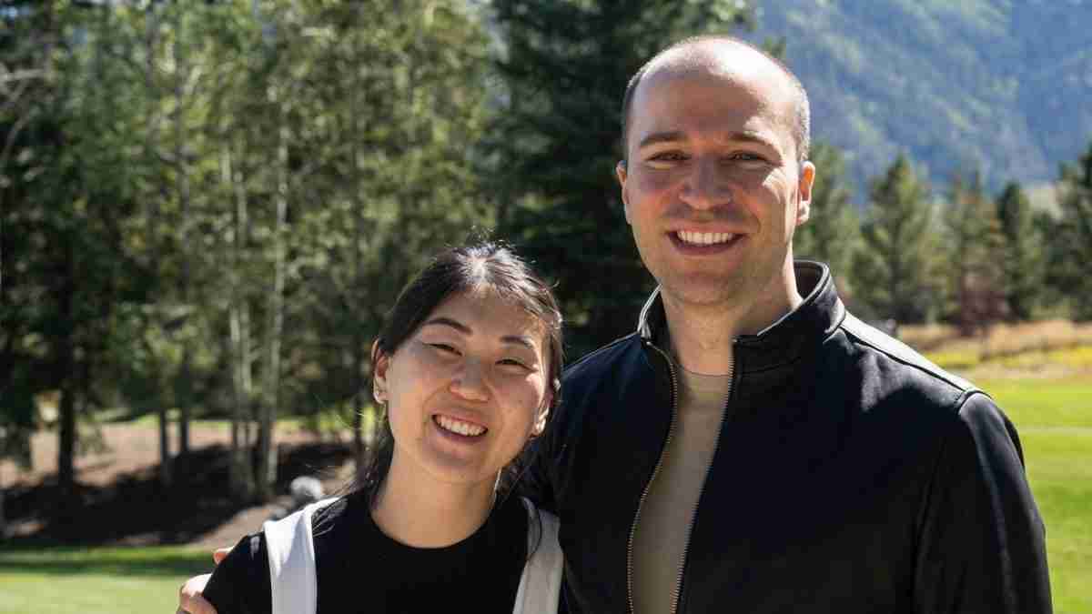 Two friends posing outdoors on a sunny day, with trees and mountains in the background, both smiling at the camera.
