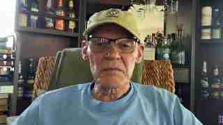 Portrait of an older man wearing a tan cap with USMC emblem and glasses, seated in a room with shelves of bottles in the background.