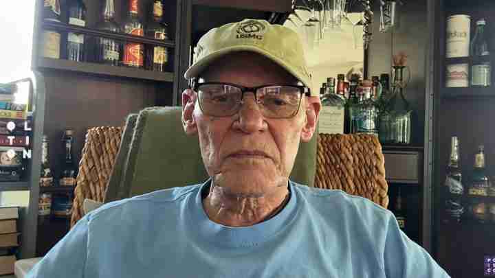 Portrait of an older man wearing a tan cap with USMC emblem and glasses, seated in a room with shelves of bottles in the background.