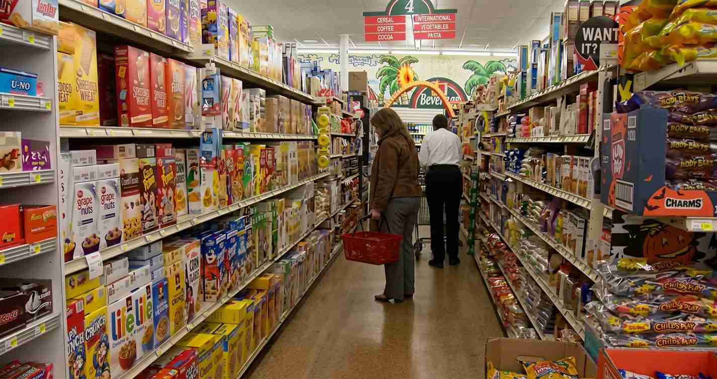 Aisle in a grocery store with cereal boxes on tall shelves; a woman in a brown jacket and a man in a white shirt stand with shopping baskets in the middle.
