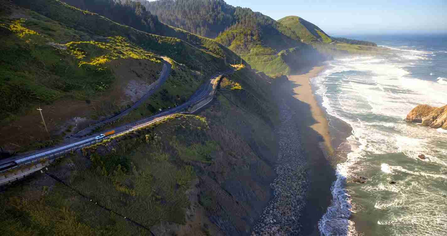 Winding coastal road along steep green cliffs overlooking the rocky shoreline and ocean. (aerial view)