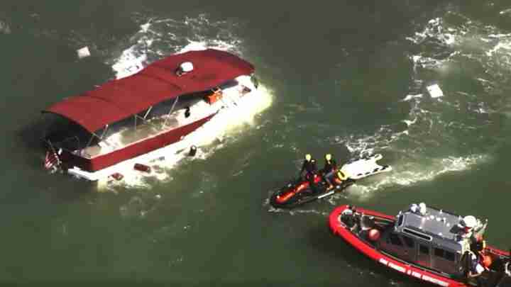 Red capsized boat with its hull partially submerged, surrounded by rescue boats and personnel in life jackets at the scene