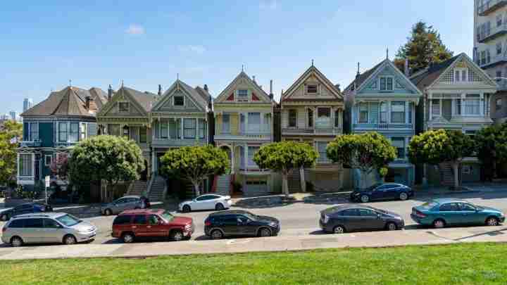 Row of pastel Victorian houses with pointy gables along a tree-lined street and parked cars in front.