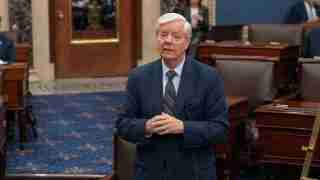 Man in a navy suit speaks in a formal legislative chamber, hands clasped in front of him.