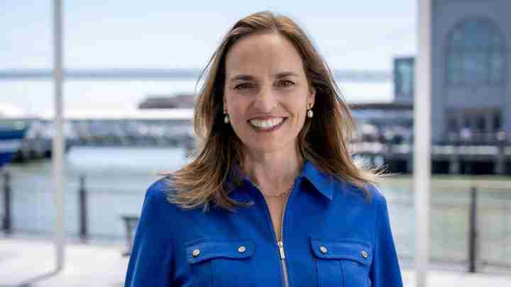 Smiling woman in a blue blouse stands at a waterfront boardwalk with boats in the background.
