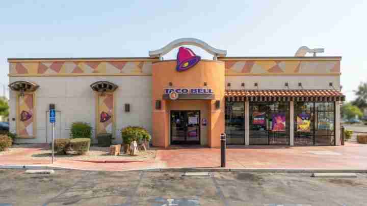 Taco Bell storefront with an orange curved entrance and purple bell logo, flanked by beige walls.