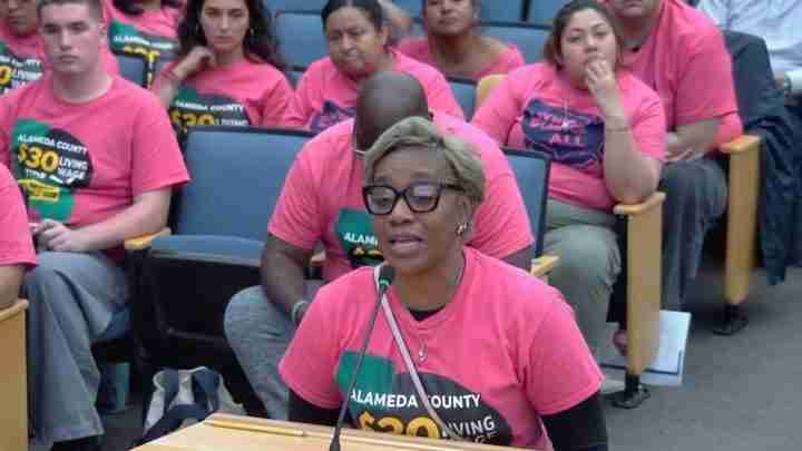 Woman with short gray hair and glasses speaks into a microphone at a podium, wearing a pink Alameda County shirts, surrounded by supporters in pink in a meeting room.