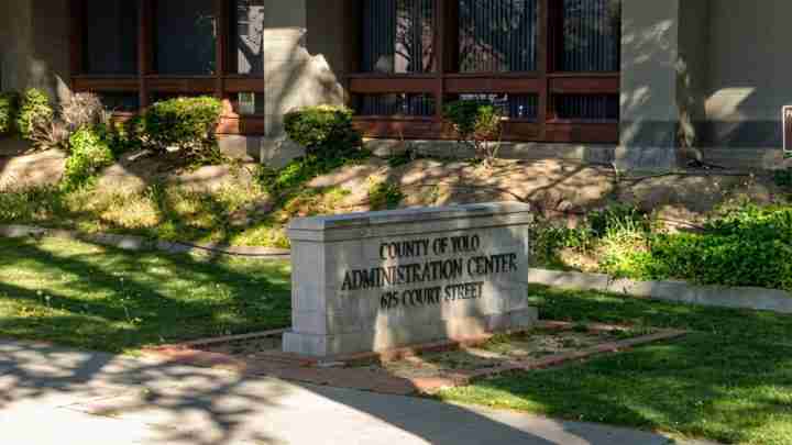 Stone pedestal sign reading 'County of Yolo Administration Center, 625 Court Street' in front of a brick-faced building with landscaping and shadows.