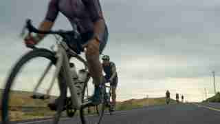 Cyclists ride on a rural two-lane road with a lead rider in the foreground and others far back under cloudy skies.