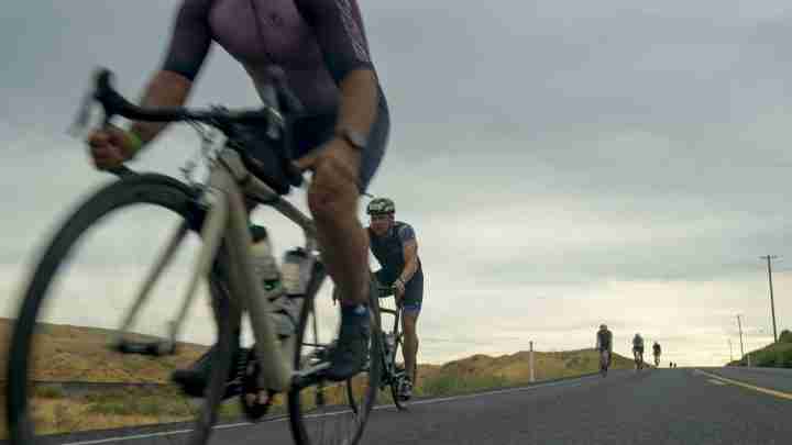 Cyclists ride on a rural two-lane road with a lead rider in the foreground and others far back under cloudy skies.