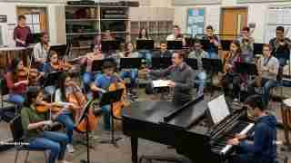 School music ensemble rehearsal in a practice room, conductor leads strings, brass, and woodwinds while a pianist accompanies at a grand piano.