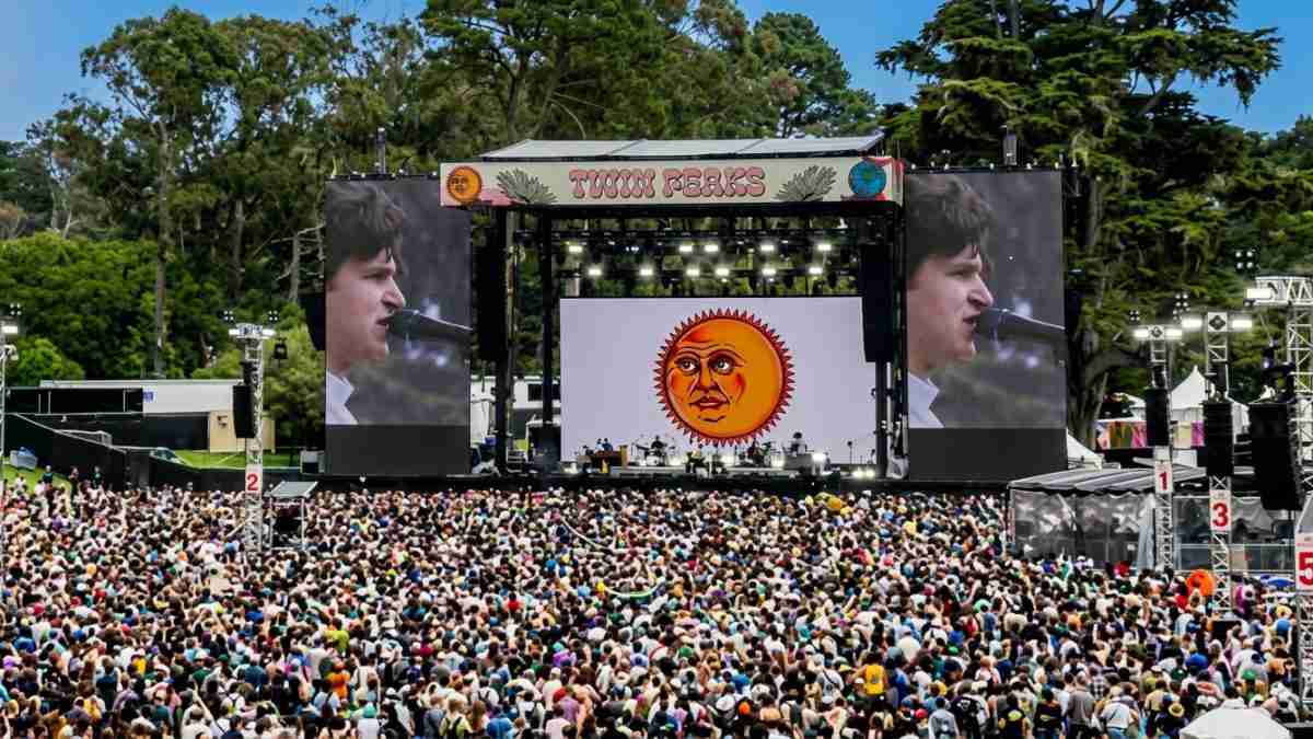 Massive audience at an outdoor concert with a sun-face logo on the stage screen.