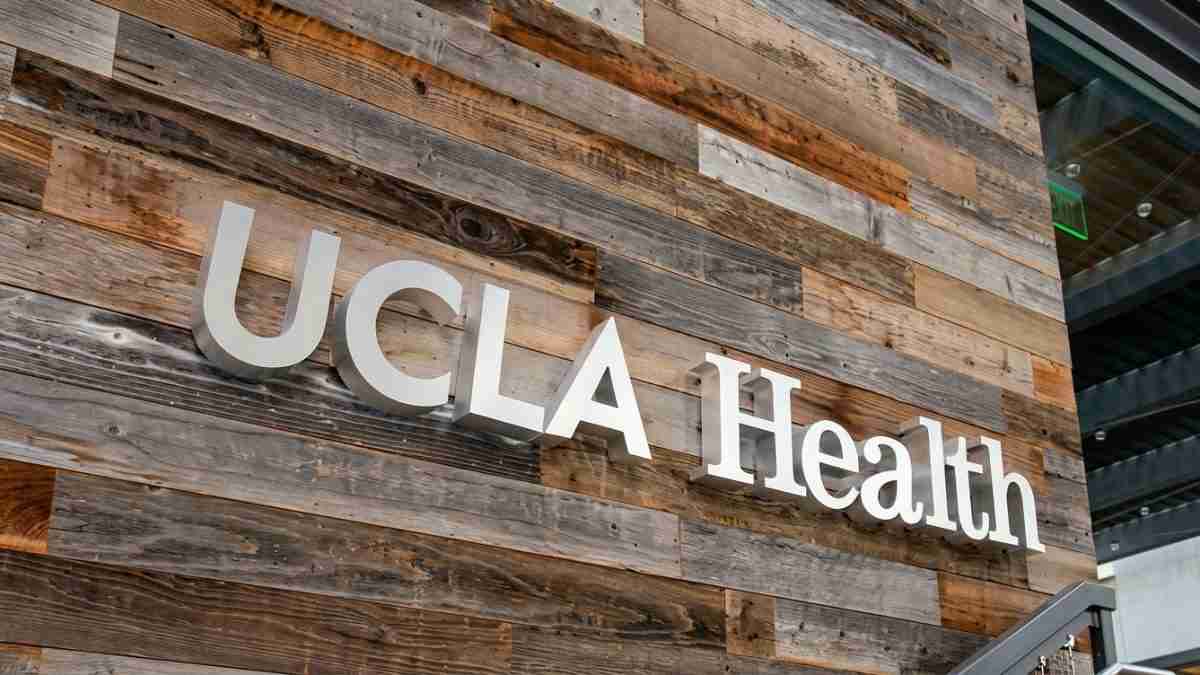 UCLA Health sign mounted on a rustic wood-paneled exterior wall near a glass entrance.