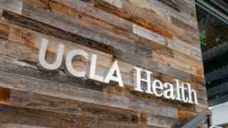 UCLA Health sign mounted on a rustic wood-paneled exterior wall near a glass entrance.