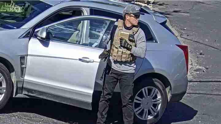 Man in a tactical vest and helmet standing beside a silver SUV with the front passenger door open, appearing on alert or in a security role