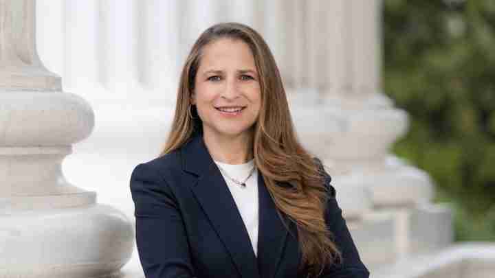 Portrait of a smiling woman with long brown hair in a navy blazer standing outdoors beside white columned architecture.